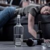 A half-empty vodka bottle sits on the floor in focus, while a drunken young woman sleeps in the background, highlighting the importance of early detection of chronic alcohol misuse.
