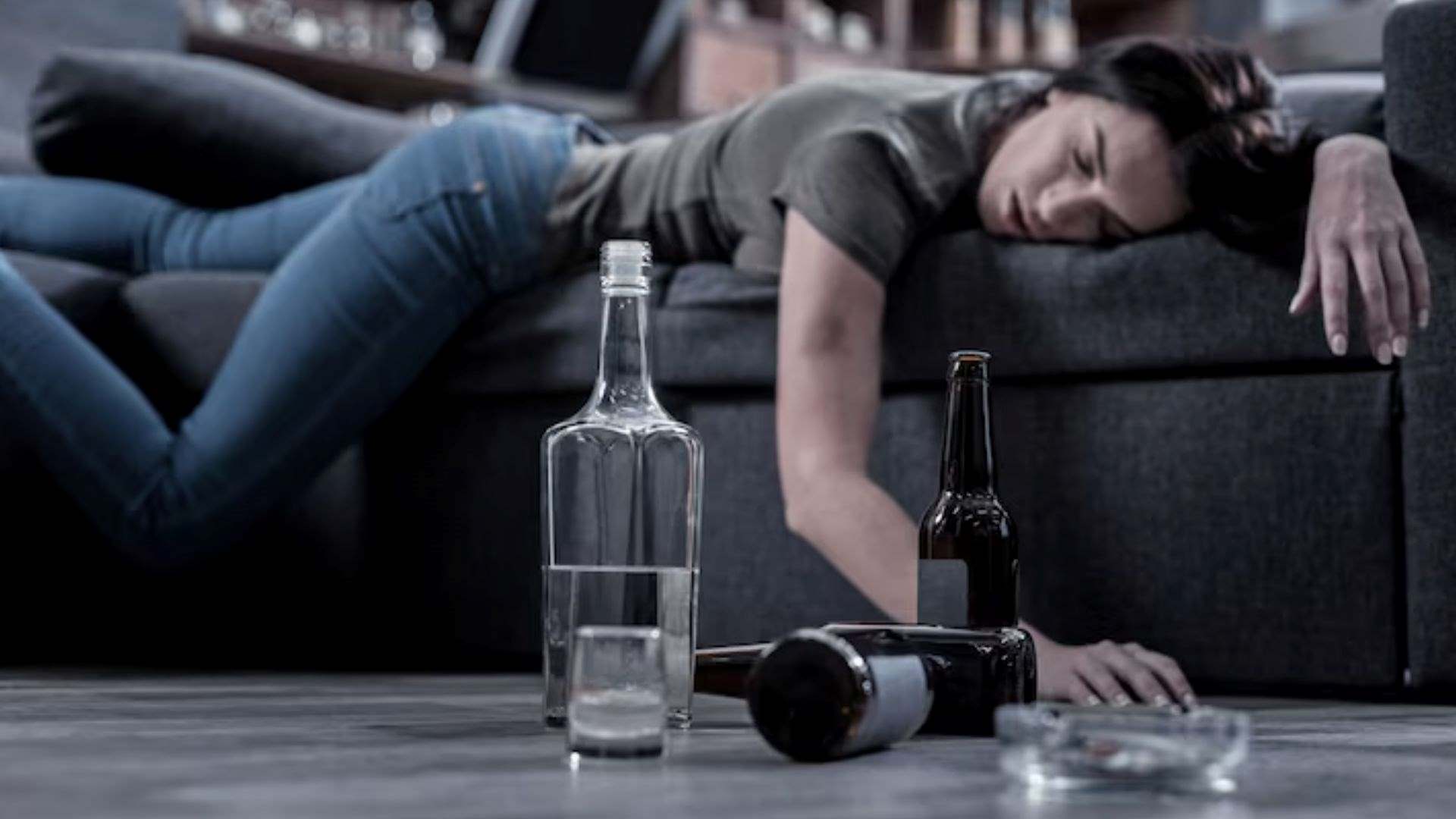 A half-empty vodka bottle sits on the floor in focus, while a drunken young woman sleeps in the background, highlighting the importance of early detection of chronic alcohol misuse.
